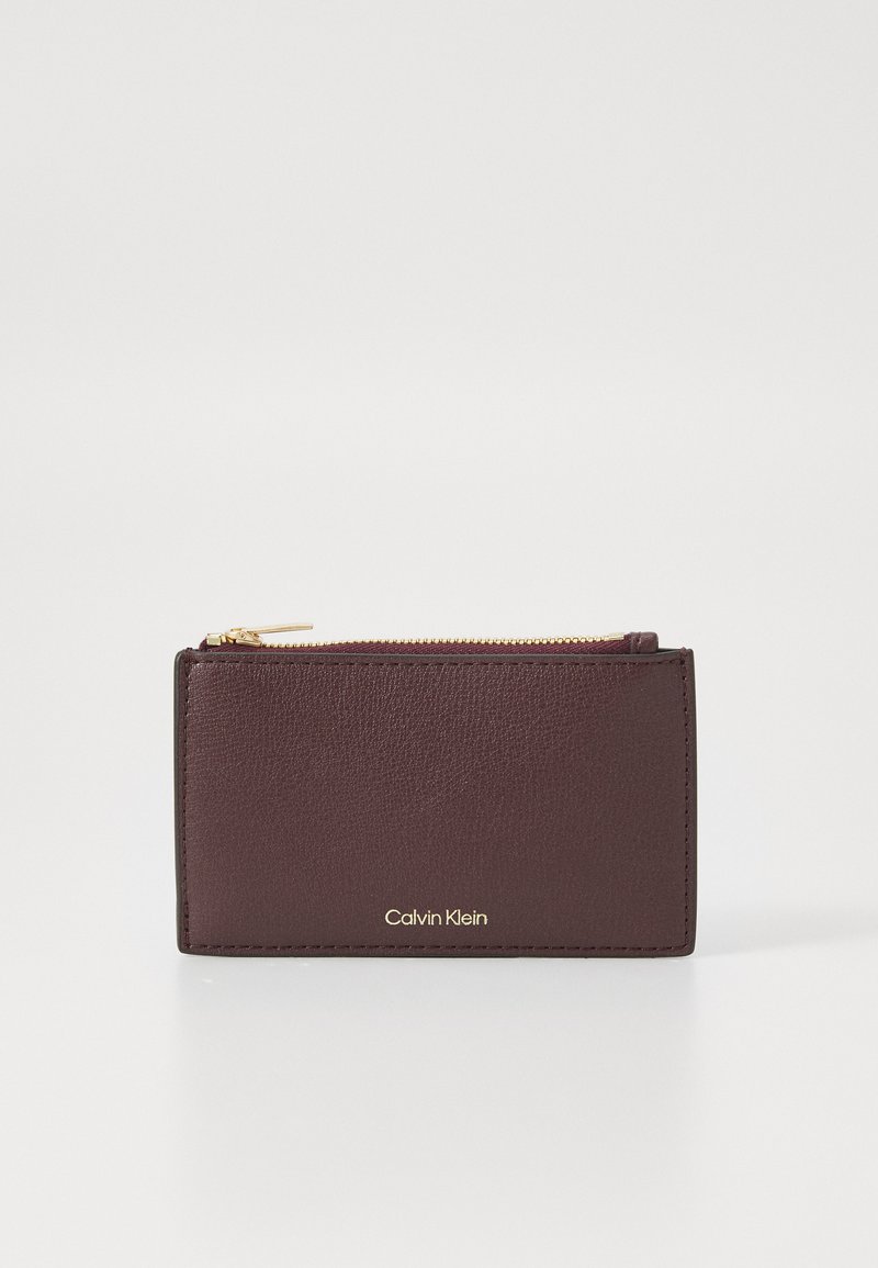 Burgundska kožna novčanik s teksturiranom površinom, zlatnim zatvaračem i urezanim logotipom "Calvin Klein" na prednjoj strani. Kompaktni pravokutni oblik.