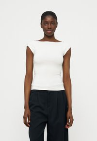 Νεαρή μαύρη γυναίκα με τα μαλλιά τραβηγμένα πίσω, φοράει εφαρμοστό λευκό κοντομάνικο τοπ και χαλαρά μαύρα παντελόνια, στέκεται μπροστά σε ουδέτερο φόντο.