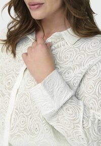 Blusa bianca a motivi con disegni a spirale, colletto con bottoni e tessuto texturizzato. Vista ravvicinata del dettaglio del tessuto e del bottone.