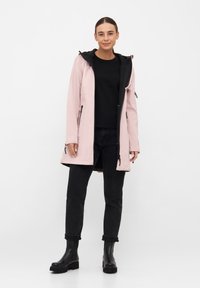 Chupa impermeable de color rosa claro con capucha negra y ribete. Presenta bolsillos con cremallera y un corte recto, combinado con pantalones negros y botas.