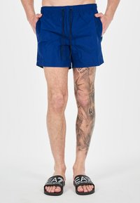 Pantaloni da bagno blu realizzati in tessuto leggero, con vita regolabile tramite cordino, tasche laterali e una texture liscia. Sandali neri con logo.