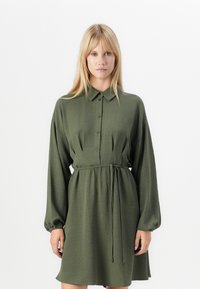 Robe chemise verte à manches longues avec un devant à boutons et une taille ceinturée, présentant un tissu texturé et une coupe décontractée.