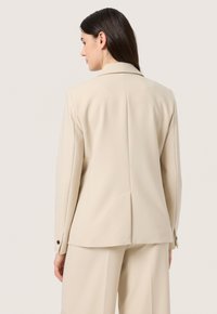 Blazer beige con una vestibilità su misura, apertura posteriore singola e polsini abbottonati. Tessuto morbido con colletto strutturato. Linee pulite e dettagli minimi.