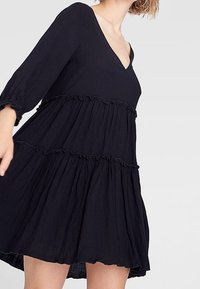Abito nero a tre strati realizzato in tessuto leggero. Presenta un collo a V e maniche lunghe con dettagli a volant lungo i vari strati.