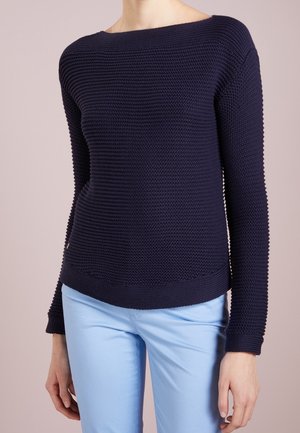 Person trägt einen marineblauen, strukturierten Strickpullover mit langen Ärmeln und hellblaue Hosen vor einem schlichten Hintergrund.