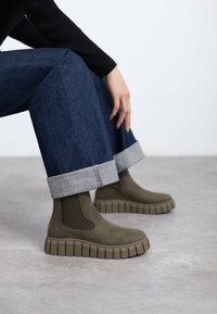 Stivali alla caviglia verde oliva con pannelli laterali elastici, suole in gomma e una piattaforma spessa e rigata. Indossati con jeans di denim risvoltati.