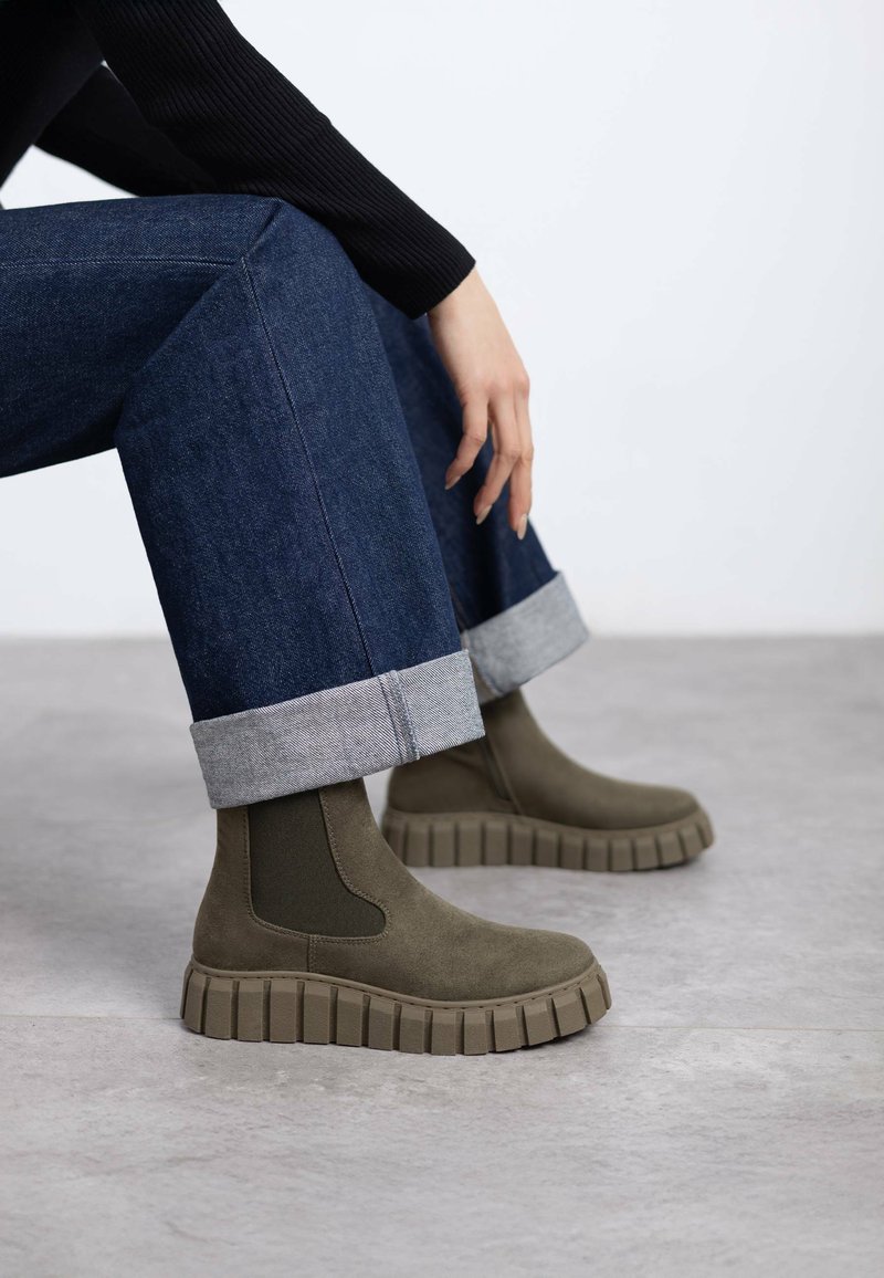 Stivali alla caviglia verde oliva con pannelli laterali elastici, suole in gomma e una piattaforma spessa e rigata. Indossati con jeans di denim risvoltati.