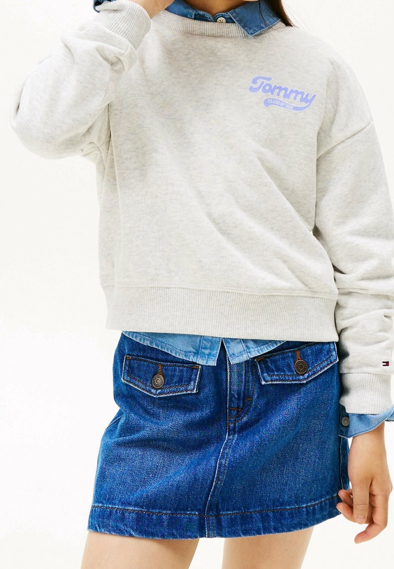 Personne portant un sweat-shirt Tommy gris clair par-dessus une chemise en denim bleu rentrée dans une jupe en denim bleu avec des poches à boutons à l'avant.