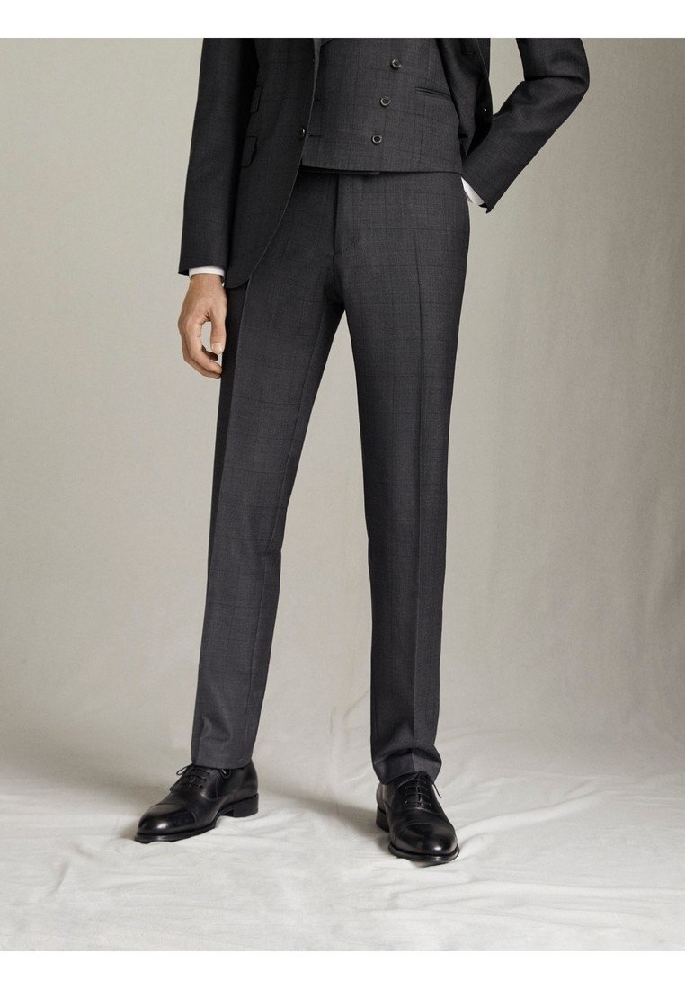 Homme portant un pantalon de costume gris foncé sur mesure, gilet assorti, chaussures habillées en cuir noir, debout avec une main dans la poche.