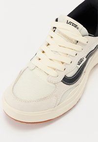 Vans sneakers with a cream upper, black side stripe, round toe, and textured mesh accents. Features cream laces and rubber outsole.