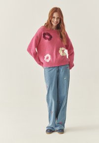 Pulover tricotat roz cu aplicații florale, asortat cu blugi largi de culoare albastru deschis, cu broderie colorată, și încălțăminte casual.