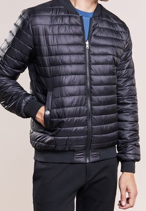Homme portant une veste bomber matelassée noire sur une chemise bleue, une main dans la poche de la veste et l'autre détendue le long du corps.
