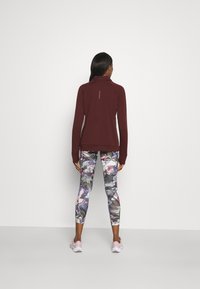 Burgunderfarbener Pullover mit langen Ärmeln und Stehkragen. Enganliegende Leggings mit einem floralen Muster in verschiedenen Rosa-, Lila- und Grautönen.