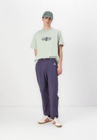 Light green graphic T-shirt with a round logo, purple elastic waist pants with accents, and beige sneakers with red stripes.