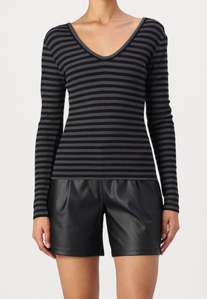 Vrouw die een zwart met donkergrijs gestreepte, lange mouw V-hals top en zwarte leren korte broek draagt, staand tegen een effen achtergrond.