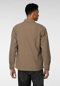 Brun arbeidsjakke med lange ermer, knapplommer, rett snitt og yoke-design. Myk stofftekstur. Brukt med mørke jeans.