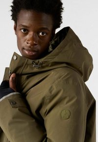 Jongere in een olijfgroene hoodie, met één hand de kraag vasthoudend, recht vooruitkijkend met een neutrale uitdrukking.