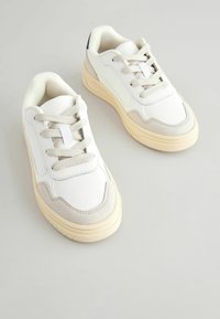 Witte synthetische sneakers met beige suède accenten, ronde neus, vetersluiting, gestructureerde tong en een dikke crème kleurige zool.