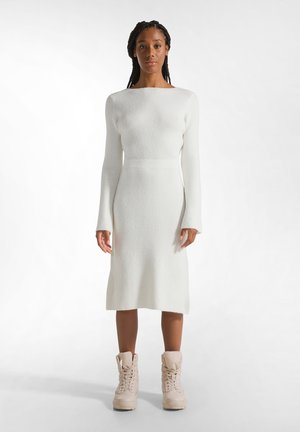Hvid ribbet strikket kjole med lange ærmer, en tætsiddende talje og midi længde; parret med lyse beige snørebåndsstøvler.