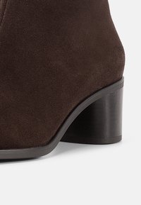 Brązowy zamszowy botek na kostkę z zaokrąglonym noskiem i czarnym, warstwowym obcasem. Gładka faktura i minimalne zdobienia podkreślają jego elegancki design.