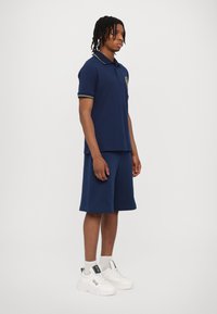 Mørkeblå polo skjorte med gule detaljer og logo, kombinert med mørkeblå shorts. Fullført med hvite sneakers og ankelsokker.