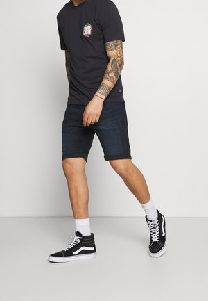 Camiseta negra con un logo, pantalones cortos de mezclilla oscura y zapatillas altas negras con acentos blancos. Tatuajes visibles en el brazo.