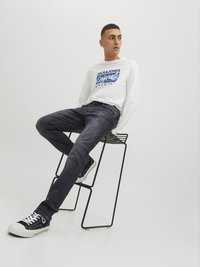 Camisa blanca de manga larga con texto gráfico azul, jeans de mezclilla oscura y zapatillas negras con suelas blancas, sentado en un taburete minimalista de metal.