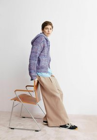 Strickkapuzenpullover in Lila- und Blautönen mit durchgehendem Reißverschluss vorne; kombiniert mit einem hellblauen Oberteil und beigen Hosen mit weitem Bein. Schwarze Sneakers auf einem Holzstuhl.