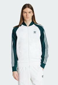 Bílá zipová sportovní bunda s zelenými akcenty a třemi bílými pruhy na rukávech. Obsahuje žebrový límec a vyšívané logo.