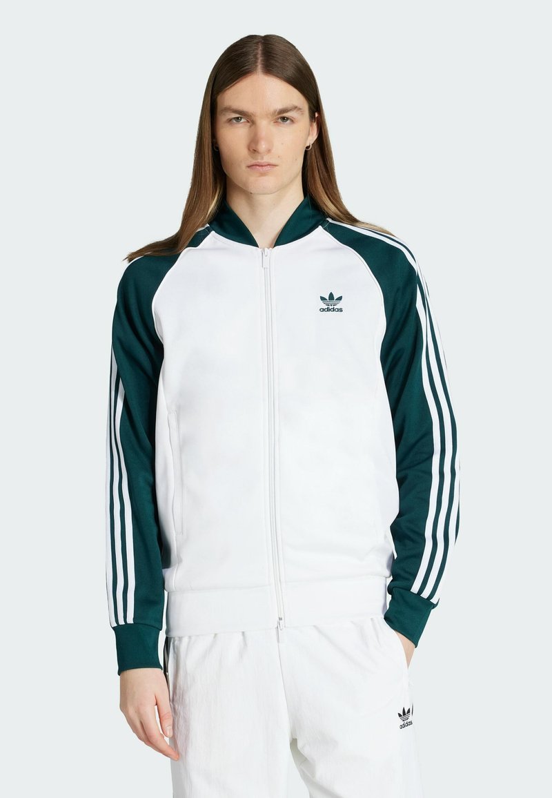 Bílá zipová sportovní bunda s zelenými akcenty a třemi bílými pruhy na rukávech. Obsahuje žebrový límec a vyšívané logo.