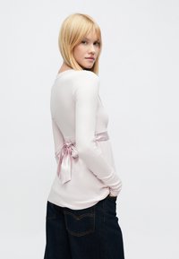 Világos rózsaszín, hosszú ujjú felső, húzott hátrésszel és selyemszalag díszítéssel. Bordázott textúrájú, sötét farmernadrággal párosítva a kontraszt érdekében.