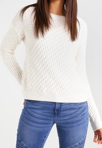 Frau trägt einen weißen strukturierten Strickpullover und blaue Jeans, steht mit einer Hand hinter ihrem Rücken vor einem schlichten Hintergrund.