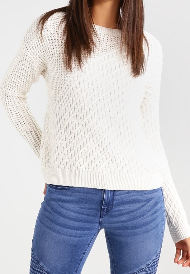 Frau trägt einen weißen strukturierten Strickpullover und blaue Jeans, steht mit einer Hand hinter ihrem Rücken vor einem schlichten Hintergrund.