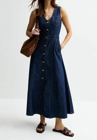 Denim maxikjol med V-ringning, ärmlös design, med gula knappar nedtill och fickor. I kombination med bruna sandaler och en väska.