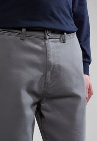 Pantaloni grigi con una trama liscia, dotati di chiusura a bottone e zip, passanti per cintura e cuciture a contrasto. Indossati con una camicia a maniche lunghe blu navy.