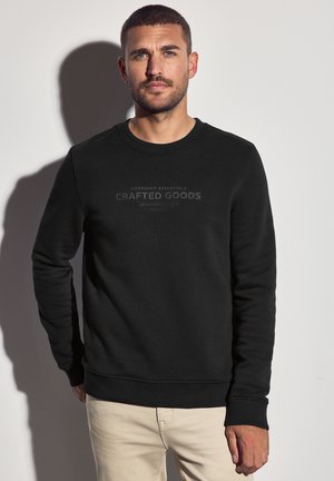 Hombre con cabello corto castaño y barba, vistiendo un sudadera negra con el texto "Crafted Goods", de pie contra una pared gris clara.