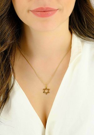 Femme aux cheveux bruns lâchés portant un collier pendentif étoile de David en or et un chemisier blanc à col en V.