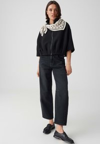 Woman standing wearing a black zip-up top, black wide-leg jeans, black loafers, and a white polka dot scarf tied around her neck.