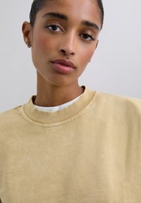 Gelber Pullover mit geripptem Rundhalsausschnitt, der eine weiche Textur und subtile Farbvariationen aufweist. Darüber liegt ein weißes Oberteil.