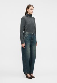 Γυναίκα με γκρι πουλόβερ ζιβάγκο, φαρδιά ξεθωριασμένα μπλε τζιν και καφέ παπούτσια με τακούνια, στεκεμένη μπροστά σε λευκό απλό φόντο.