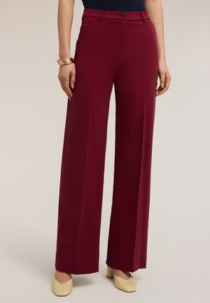 Szerokie bordowe spodnie noszone z beżowymi butami na słupku i ciemnym topem, dopełnione złotymi bransoletkami na nadgarstku.