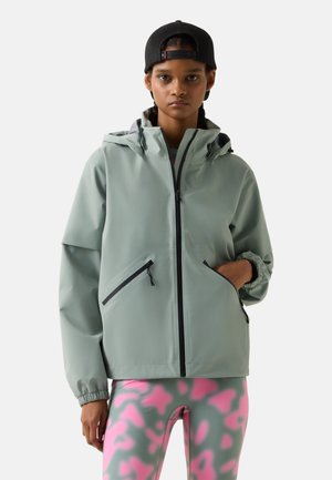 Young person wearing a light gray hooded jacket, black backward cap, and pink and gray patterned leggings, standing against a plain background.