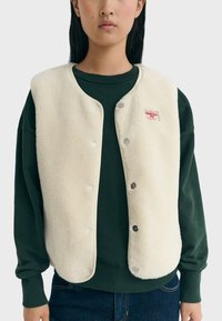 Cremefarvet fleecevest med blød tekstur, trykknaplukning og et lille rødt mærke; båret over en mørkegrøn sweatshirt.