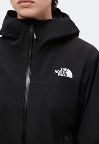 Personne portant une veste North Face noire avec une capuche et une fermeture éclair frontale remontée jusqu'au cou.