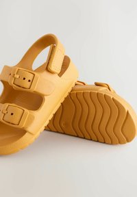 Gelbe Kunststoffsandalen mit drei verstellbaren Riemen und Schnallen, strukturierter Oberseite und einer welligen Musterung der Sohle für besseren Halt.