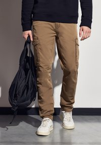 Khaki cargo kalhoty se postranními kapsami, vyrobené z bavlněné směsi. Nosené s černou mikinou a bílými teniskami, držící černý batoh.