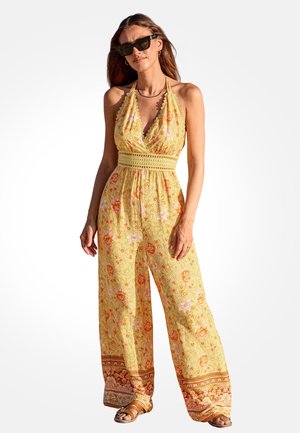 Frau mit Sonnenbrille trägt einen gelben, geblümten Jumpsuit mit Neckholder, weitem Bein und gemustertem Saum, steht vor weißem Hintergrund.
