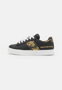 Black leather sneaker featuring golden floral patterns, white laces, and a textured rubber sole with "VERSACE JEANS COUTURE" branding.