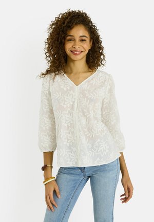 Mujer sonriente con cabello rizado que lleva una blusa blanca bordada con flores, de manga tres cuartos, y vaqueros azul claro.