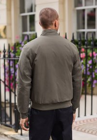 Khakifarbene Bomberjacke aus leichtem, glattem Stoff, mit gerippten Bündchen und einem engen Saum. Kombiniert mit dunkler Hose, Ansicht von hinten.